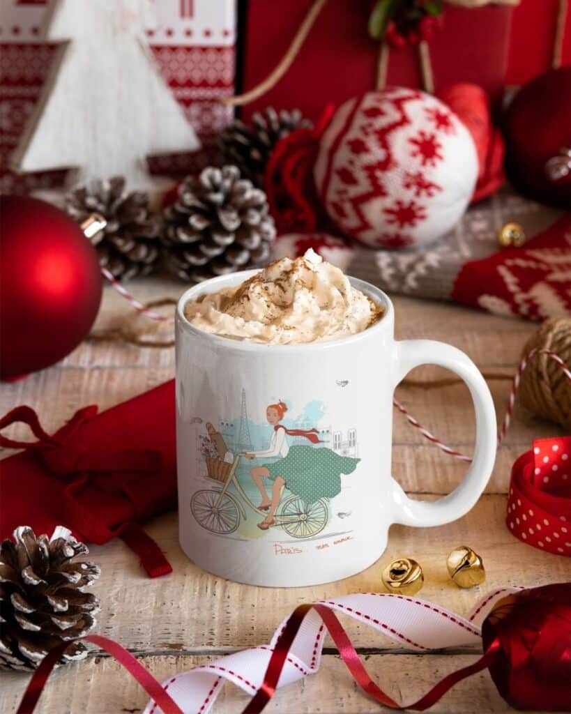  A white mug adorned with an illustration of a woman riding a bicycle in Paris, filled with whipped cream-topped hot chocolate. The scene includes festive red and white decorations, pine cones, and ribbons, evoking a cozy holiday spirit.