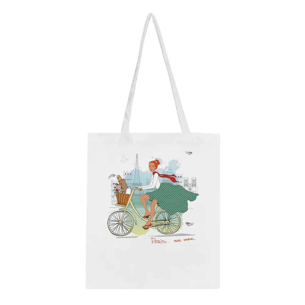 A white tote bag featuring an illustration of a stylish woman riding a bicycle with the Eiffel Tower and Parisian skyline in the background, along with the words "Paris mon amour," exuding a chic, whimsical Parisian charm.