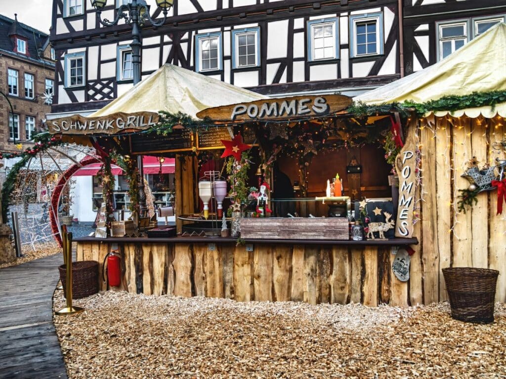 A charming wooden Christmas market stall with signs reading "Schwenk Grill" and "Pommes," decorated with festive lights, garlands, and holiday ornaments, set against a traditional half-timbered building.