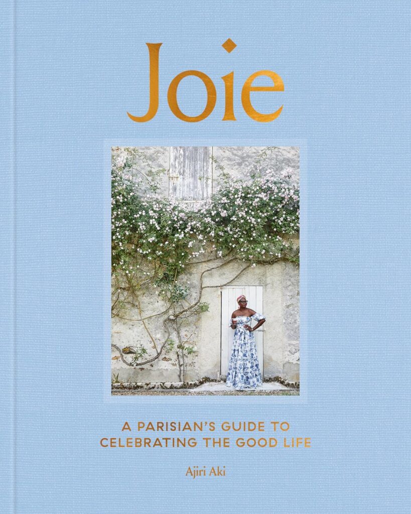 Joie: A Parisian's Guide to Celebrating the Good Life by Ajiri Aki showcases a serene, rustic setting with a woman in a floral dress standing before a stone wall covered in flowering vines. The cover suggests a tranquil, luxurious approach to enjoying the simple pleasures of Parisian life.
