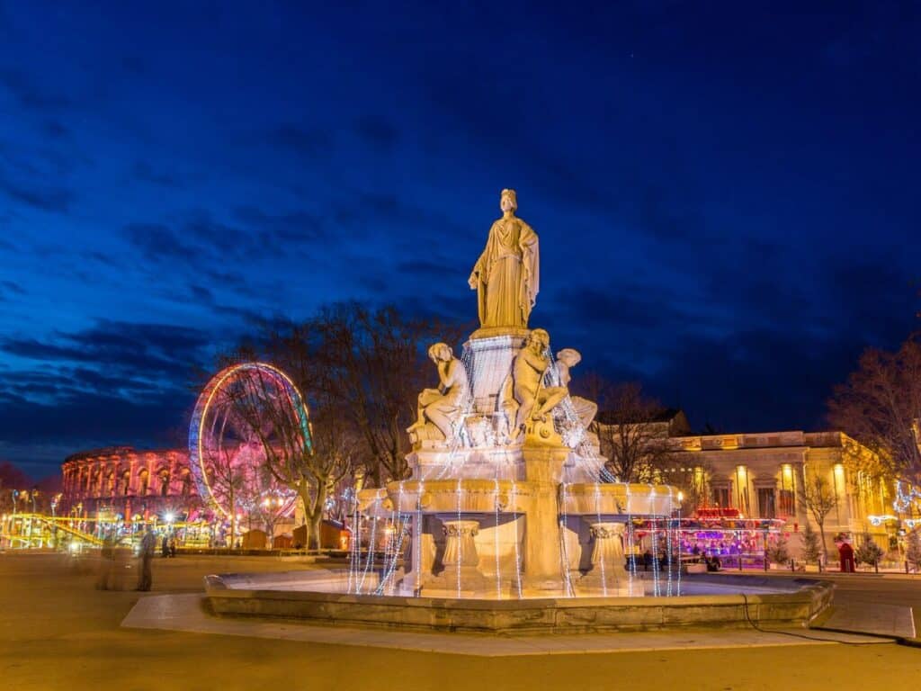 A nighttime view of a grand fountain statue in Nîmes, adorned with holiday lights, with a colorful Ferris wheel and market stalls glowing in the background under a dark blue sky.