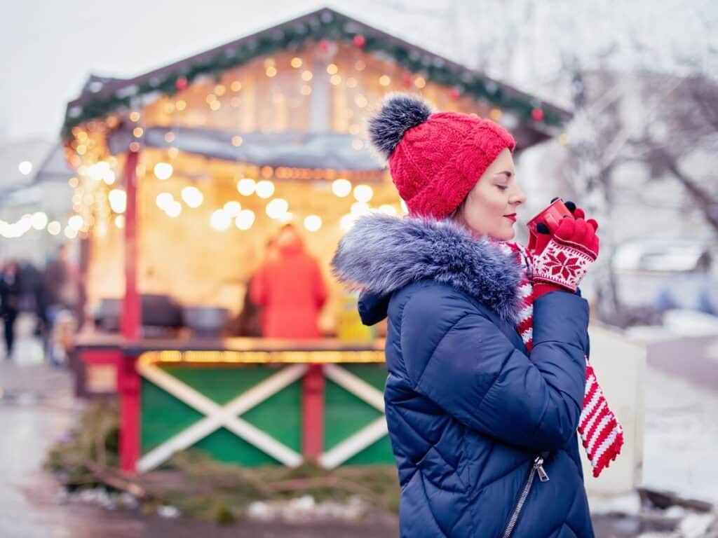 A woman in a red knit hat and gloves, bundled in a blue winter coat with a faux fur-lined hood, enjoys a hot drink against a softly blurred background of a festive holiday market booth adorned with warm lights.