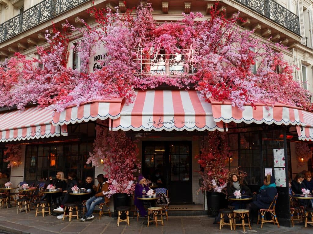 The exterior of a café in Paris with red and white striped awnings adorned with lush pink blossoms, bustling with customers enjoying outdoor dining.