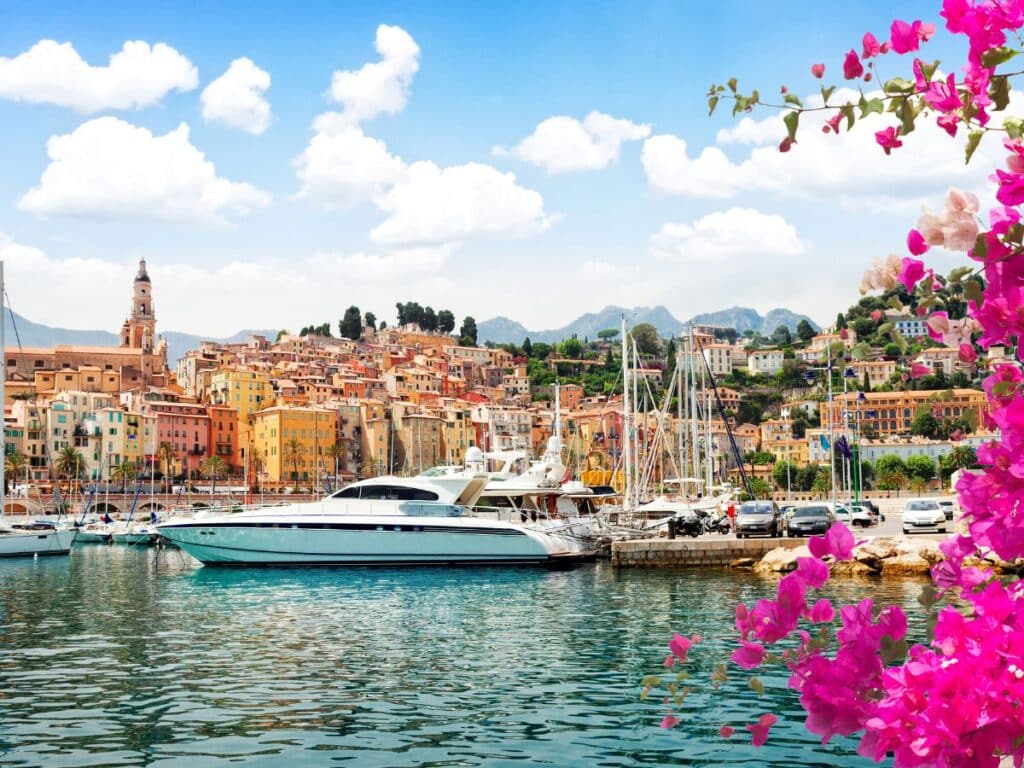 Vibrant harbor of Menton on the French Riviera with colorful hillside buildings, yachts docked at the marina, and bright pink bougainvillea framing the scene. A must-visit stop on a French Riviera road trip.