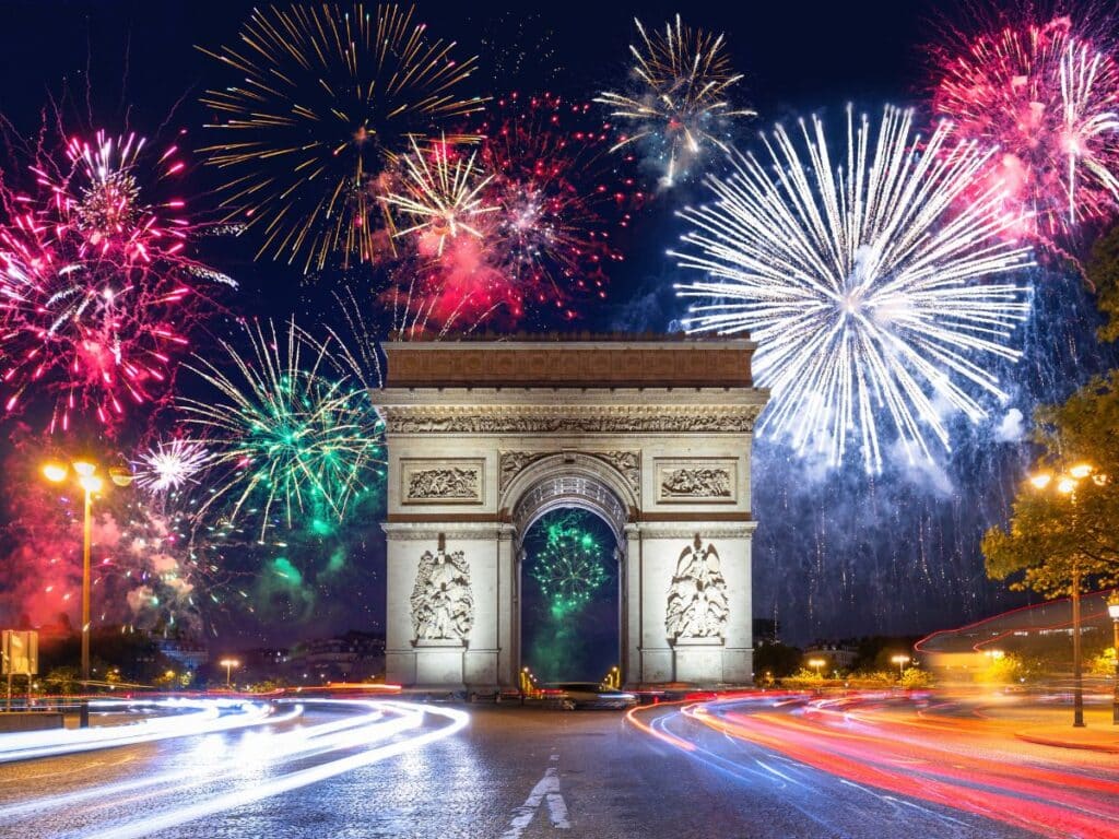 A vibrant display of colorful fireworks exploding in the night sky above the Arc de Triomphe in Paris, with light trails from cars creating dynamic movement in the foreground.