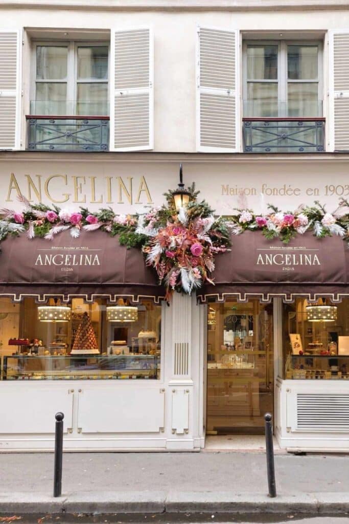 The exterior of "Angelina," a historic Paris café, featuring elegant white and brown décor with floral garlands and window displays.