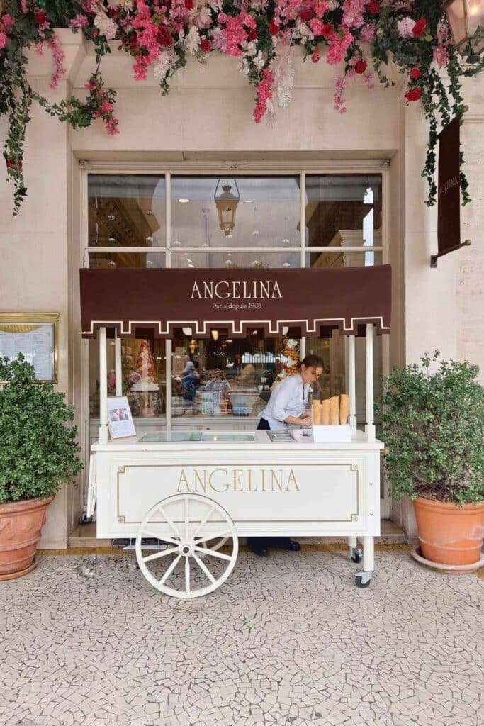 A charming Angelina café cart with a brown awning, serving ice cream and pastries, surrounded by floral decorations and greenery.