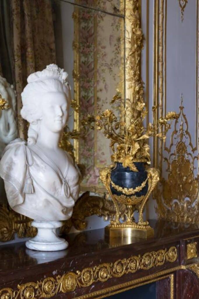 A marble bust of a woman, possibly Marie Antoinette, sits on an ornate marble surface beside an intricately decorated gold vase filled with gilded flowers, reflecting opulence in a historical setting.