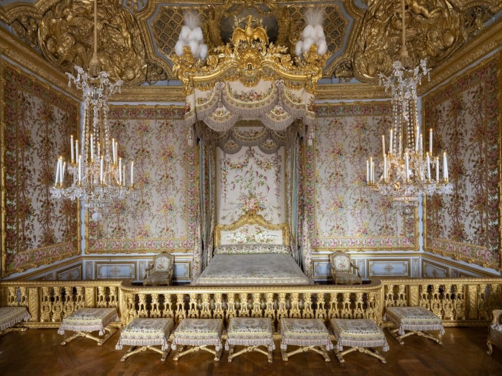 A lavish bedroom with a gilded bed draped in floral-patterned curtains and wallpaper, featuring chandeliers, upholstered stools, and elaborate gold detailing, characteristic of Versailles-style luxury.