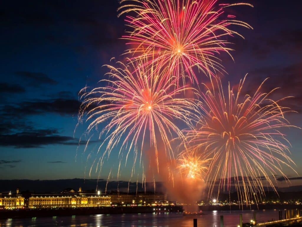 Bright golden and pink fireworks illuminate the night sky above Bordeaux, reflecting beautifully on the calm water of the Garonne River with historic buildings glowing in the background.