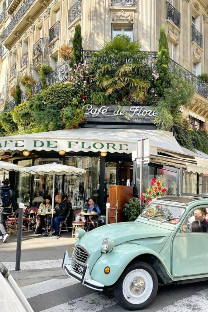 The lush, green exterior of Café de Flore in Paris, adorned with flowers and plants, with outdoor seating and a vintage mint green car parked nearby.