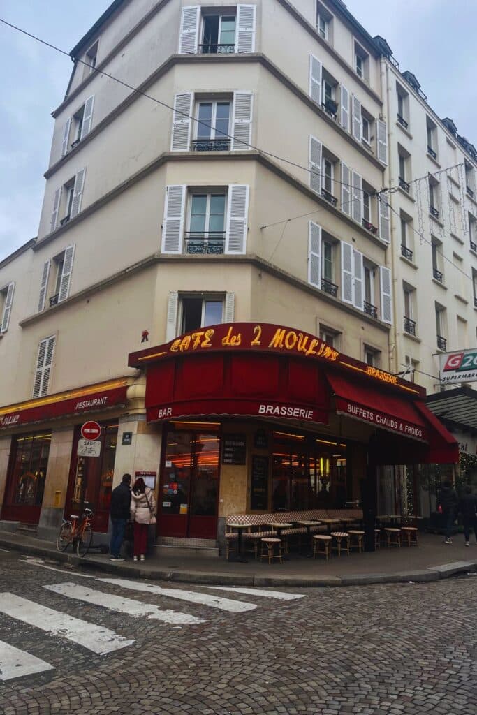 The exterior of Café des Deux Moulins in Paris, featuring red awnings and outdoor seating on a cobbled street, a café made famous by the movie Amélie.