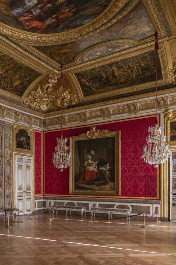 A richly decorated room with red damask walls, two crystal chandeliers, gold moldings, and a large framed painting, emphasizing the grandeur of the palace's interior.