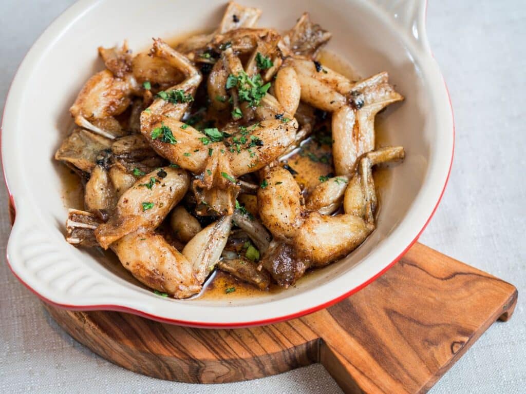 A close-up of a dish featuring golden-brown cooked frog legs garnished with fresh herbs, served in a rustic ceramic dish on a wooden board.