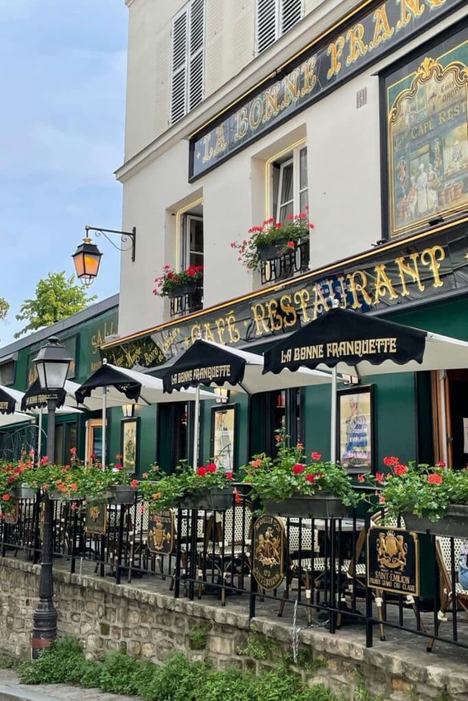 La Bonne Franquette café in Paris, showcasing a charming green façade, gold signage, and red flowers adorning the outdoor terrace.
