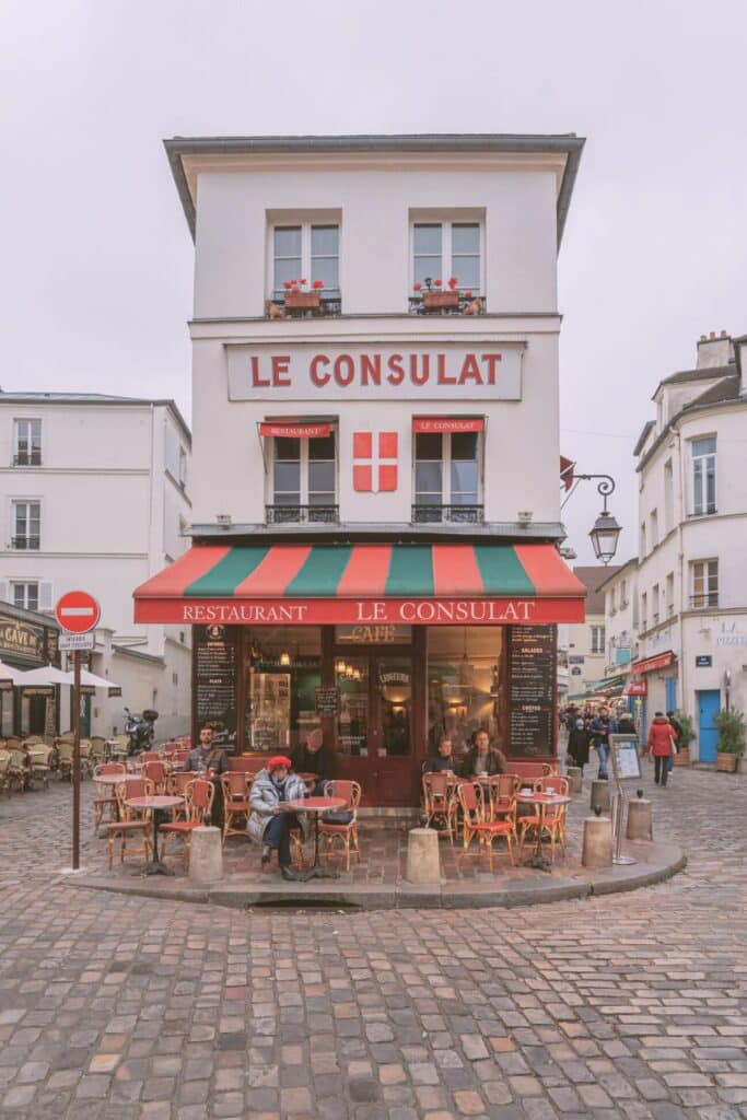 Le Consulat café in Montmartre, with red and green awnings, a white façade, and outdoor seating on a picturesque cobblestone street.