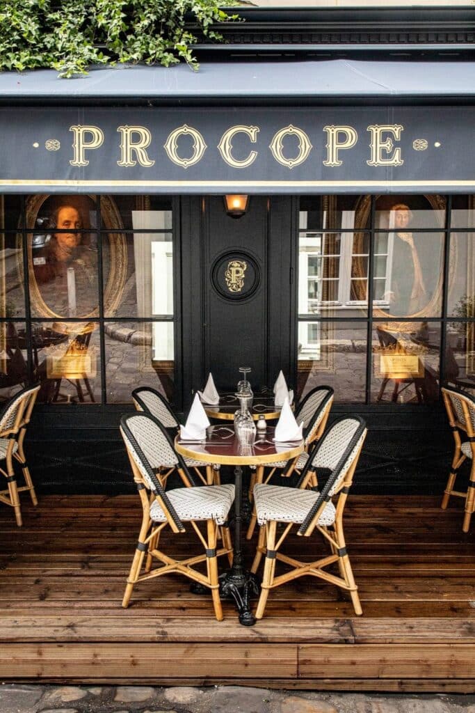 The exterior of Le Procope café, with elegant black signage, vintage portraits in windows, and outdoor seating set with fine tableware.