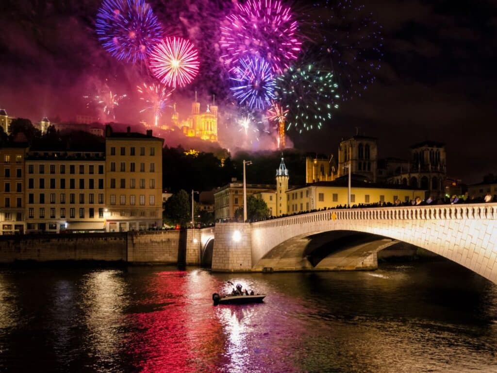 A vivid array of fireworks in shades of pink, purple, and blue lights up the sky over Lyon, with a brightly lit bridge and river below, and historic buildings perched on a hill in the distance.