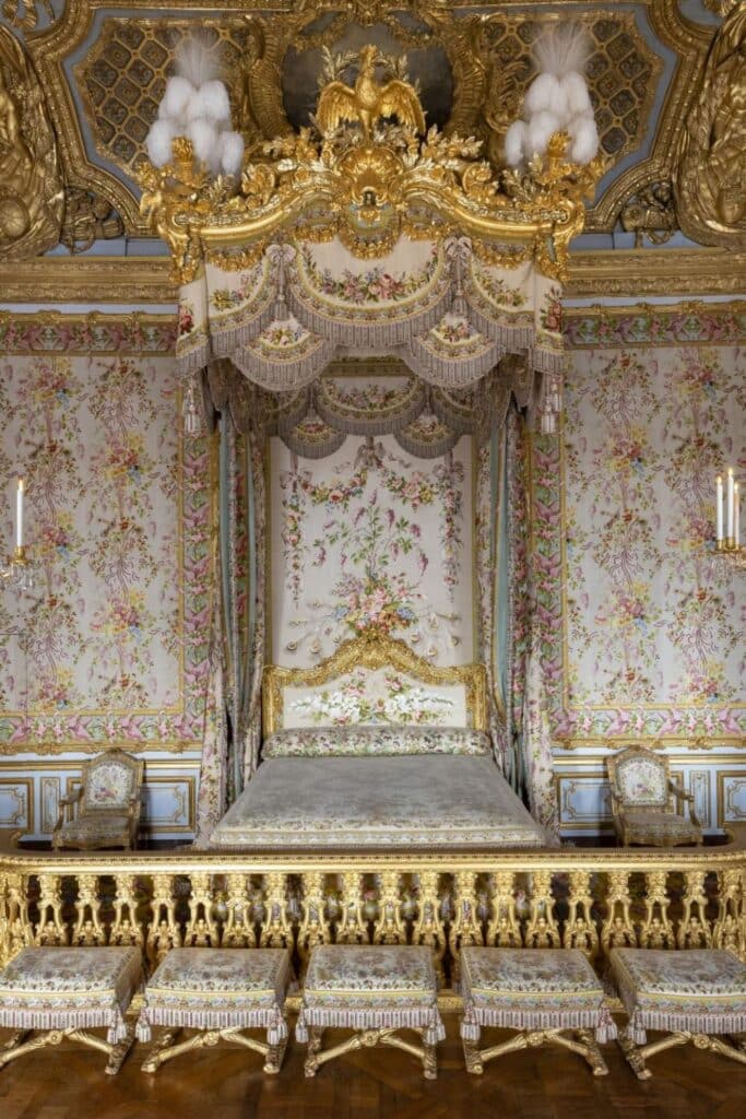 A gilded canopy bed draped with floral fabric and surrounded by richly patterned wallpaper, detailed carvings, and an ornate ceiling at the Palace of Versailles.