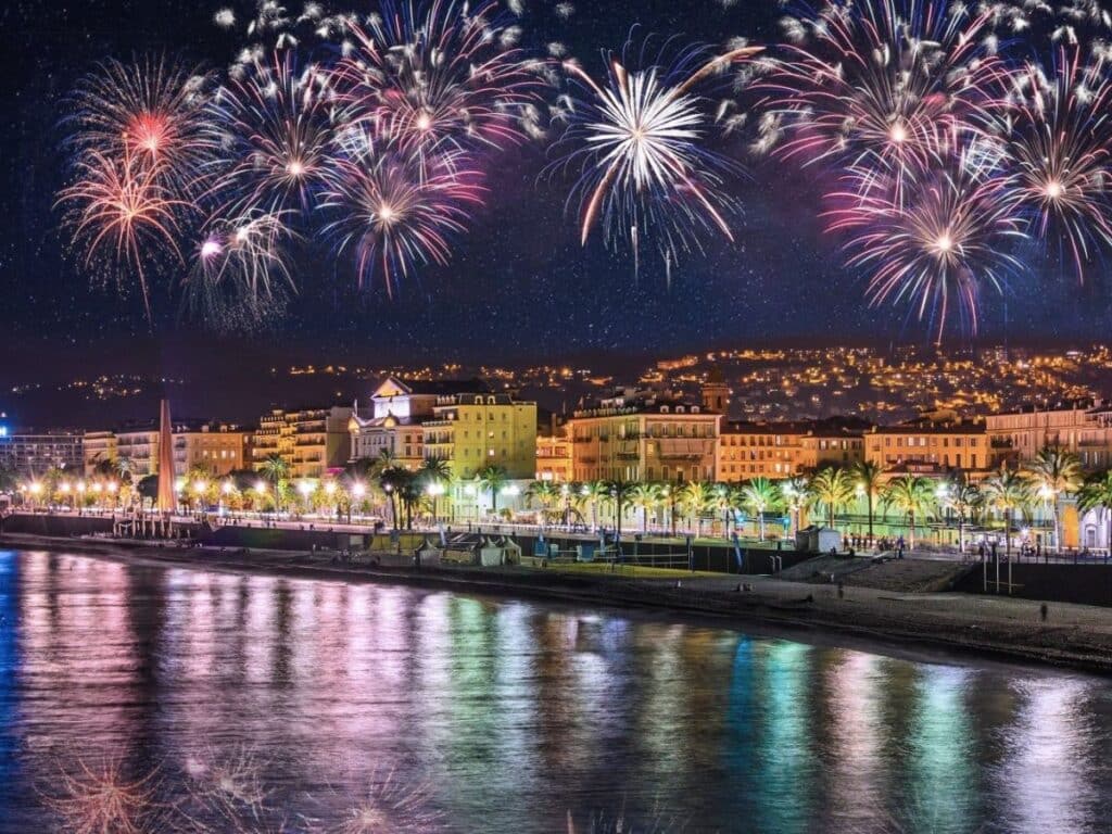 A spectacular fireworks display casts colorful reflections on the waterfront in Marseille, with the city’s promenade lined with illuminated palm trees and historic architecture in view.
