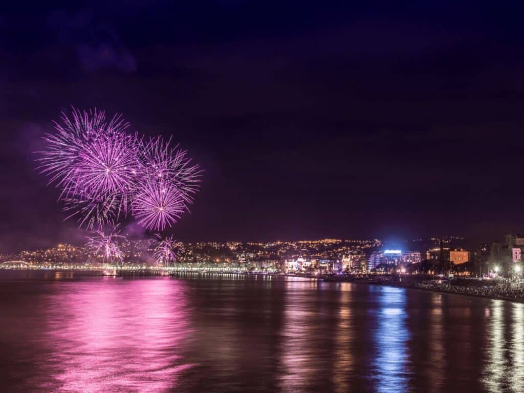 A dazzling burst of multicolored fireworks lights up the night sky above Nice, with reflections shimmering in the water and the cityscape glowing warmly in the background.