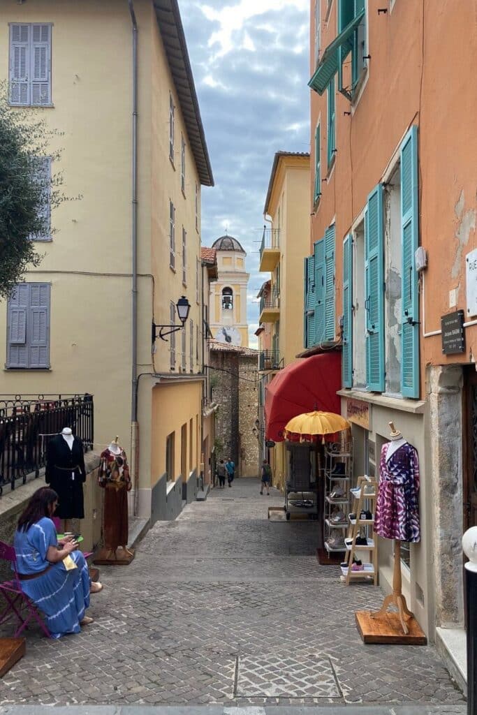 A lively scene on Rue du Poilu, Villefranche-sur-Mer, featuring boutique shops, pastel shutters, and a quaint clock tower in the background.