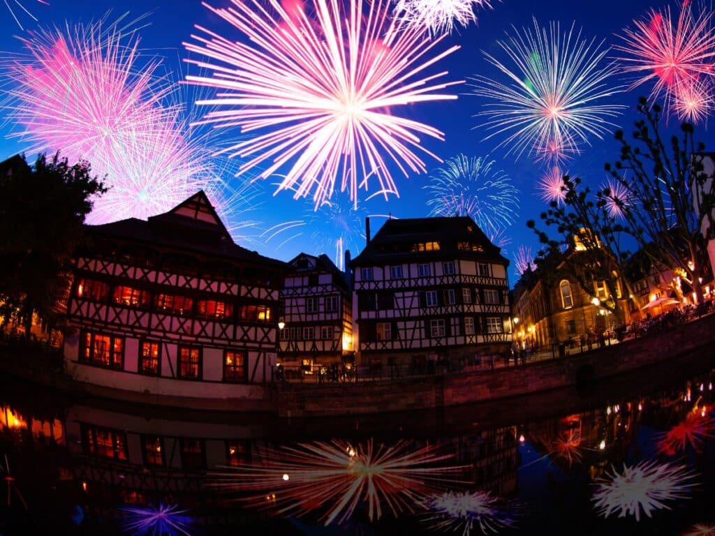 Colorful fireworks explode in the starry night sky above Strasbourg’s traditional half-timbered buildings, with their glowing windows reflected in a calm canal.