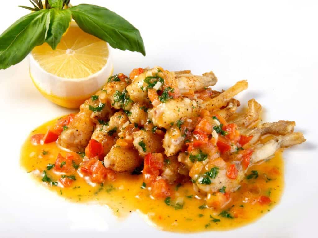 Frog legs plated with a vibrant orange and red sauce, garnished with fresh herbs, chopped tomatoes, and served alongside a decorative lemon half and basil leaves.