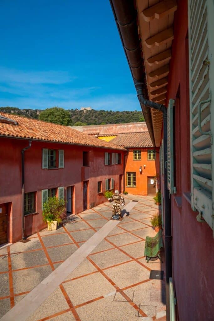 A picturesque courtyard in Villefranche-sur-Mer, featuring terracotta-tiled roofs, vivid red walls, and a unique art sculpture, framed by the surrounding hills.