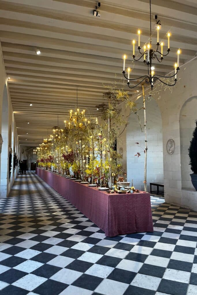 A long gallery with a black-and-white checkered floor, decorated for the holidays with chandeliers and festive floral arrangements. The arched windows and candlelight enhance the historic ambiance.