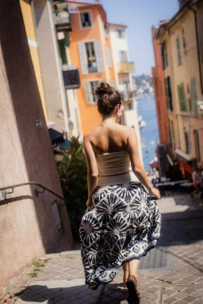 A candid shot of a woman in a flowing dress walking down a cobblestone street lined with colorful homes, with a glimpse of the bay in the background—a quintessential Instagram scene in Villefranche-sur-Mer.