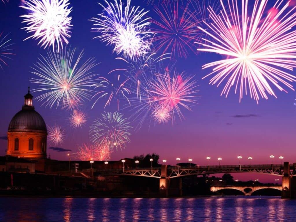 Bright pink and purple fireworks burst in the night sky over Toulouse, with the city’s iconic dome and bridge illuminated against a vibrant twilight backdrop.