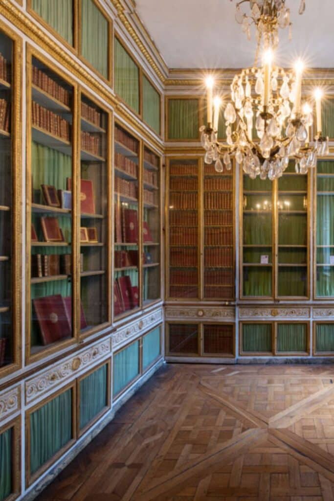 A historical library with tall bookshelves filled with leather-bound books, green curtains, gold accents, and a sparkling chandelier hanging from the ceiling.
