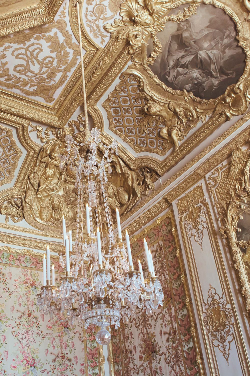 A close-up of an ornate gold ceiling adorned with intricate carvings, paintings, and a grand crystal chandelier, evoking Baroque architectural splendor.