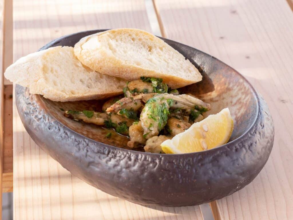 A rustic dish of cooked frog legs garnished with parsley, accompanied by slices of crusty bread and a lemon wedge, placed on a wooden table.