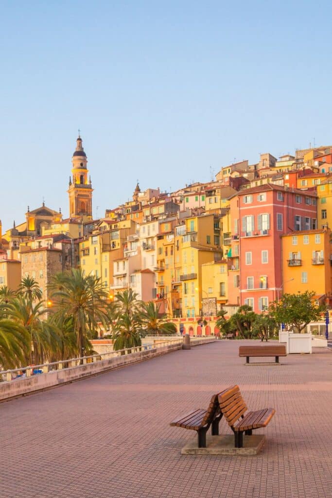 A charming walkway lined with benches and palm trees, offering a stunning view of the colorful architecture of Menton, France, under a soft evening glow.