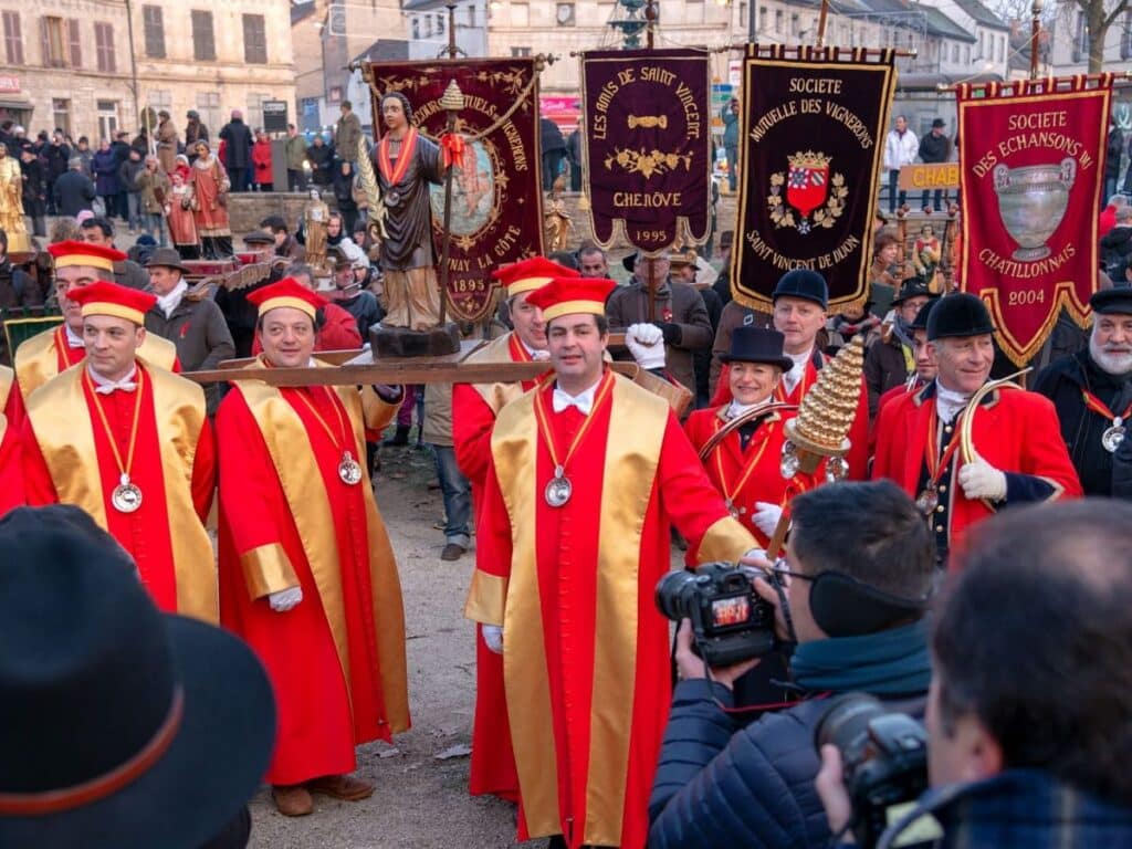 Participants dressed in traditional attire carry banners and statues during the Fête de Saint-Vincent Tournante, a vibrant January wine festival in France.