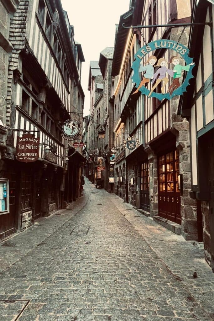 A narrow cobblestone street on Mont Saint Michel, flanked by medieval half-timbered buildings with rustic shop signs, leading up toward the island’s historic abbey.