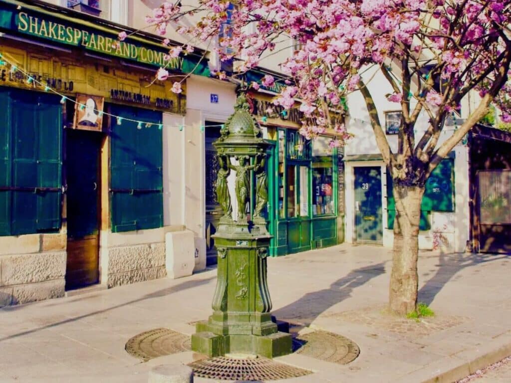 A Wallace Fountain in front of the Shakespeare and Company bookstore in Paris, framed by a pink blossoming tree. The historic green cast-iron fountain blends into the literary charm of the location.