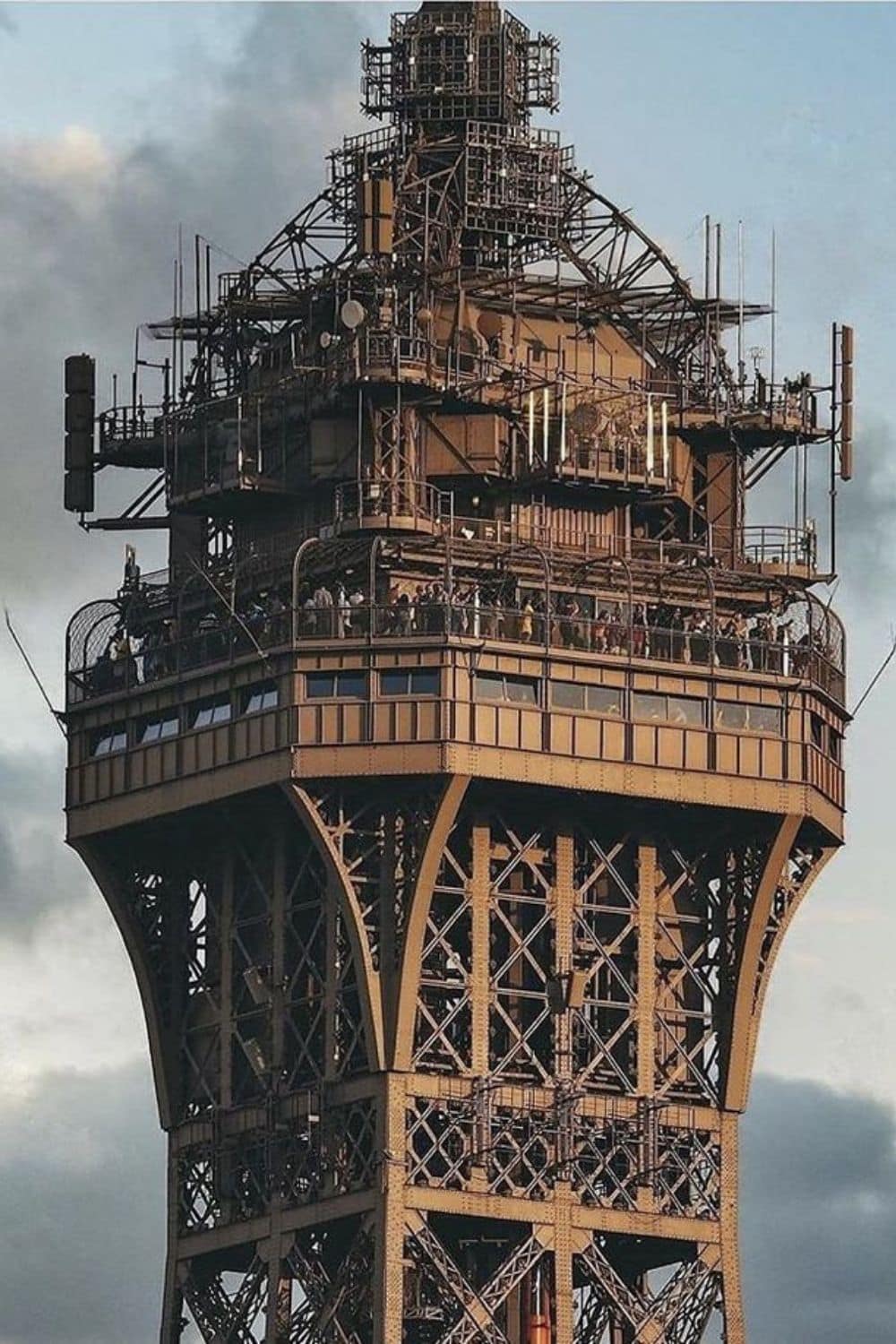 A detailed close-up of the top section of the Eiffel Tower, showcasing its intricate iron framework, observation deck, and communication equipment. The image highlights the location of the secret Eiffel Tower Apartment, once used by Gustave Eiffel as a private retreat.