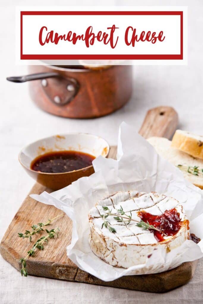 A rustic wooden board with a round of camembert cheese topped with fresh thyme and red jam, served alongside a bowl of dipping sauce and bread slices.