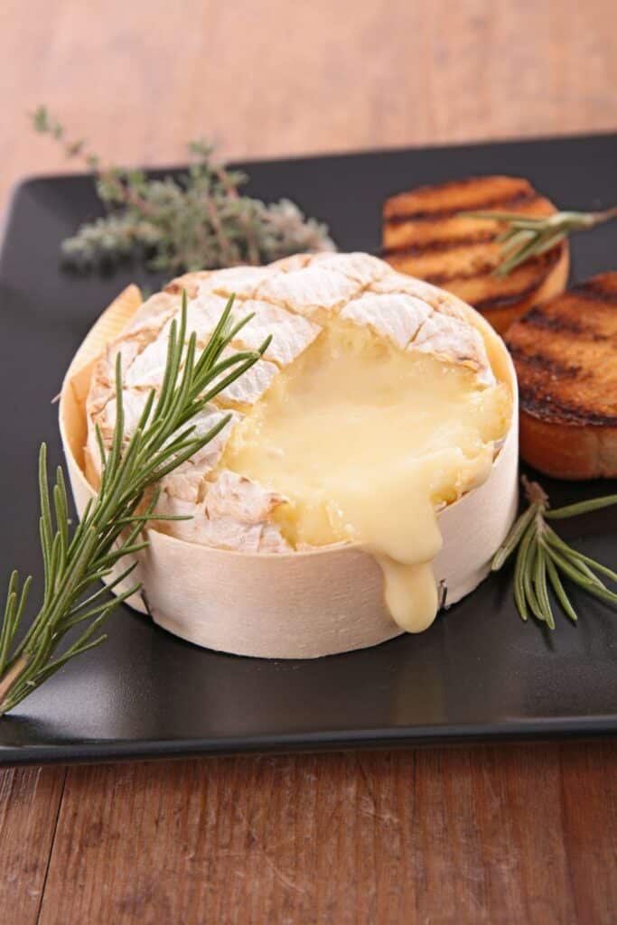 A creamy, melted camembert cheese round garnished with rosemary, served on a black plate with grilled bread slices, perfect for a savory appetizer.