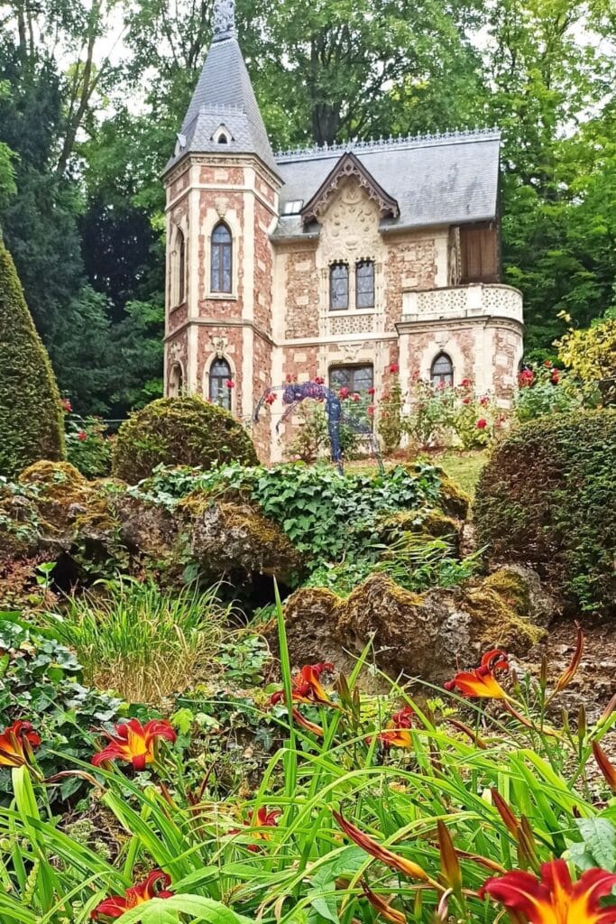 Gothic-style pavilion at Château de Monte-Cristo surrounded by lush greenery and vibrant red flowers, with pointed arches, decorative stonework, and a quaint balcony nestled among manicured gardens.