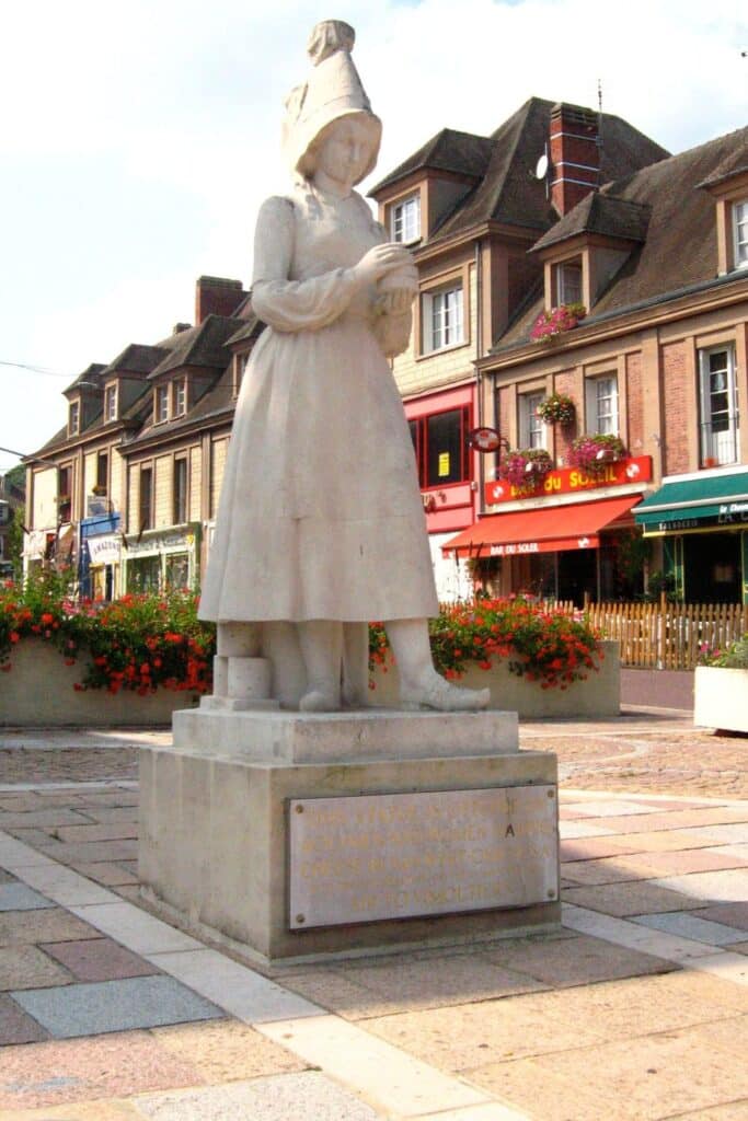 A marble statue of Marie Harel, the inventor of camembert cheese, standing in a French town square surrounded by colorful shops and flowers.