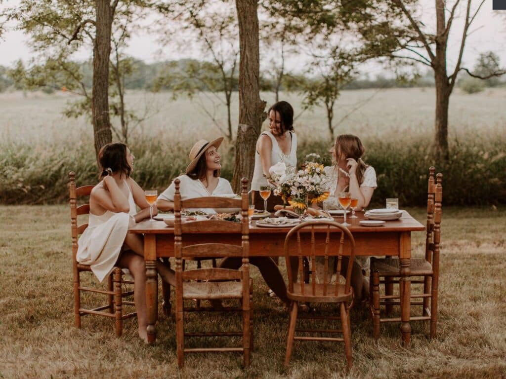Four women gather around a rustic wooden dining table set outdoors in a countryside setting, enjoying a meal with wine glasses and fresh flowers. Dressed in light, summery outfits, they laugh and converse under the shade of tall trees, with open fields stretching in the background. A dreamy representation of the idyllic countryside lifestyle, evoking the charm of moving to rural France.
