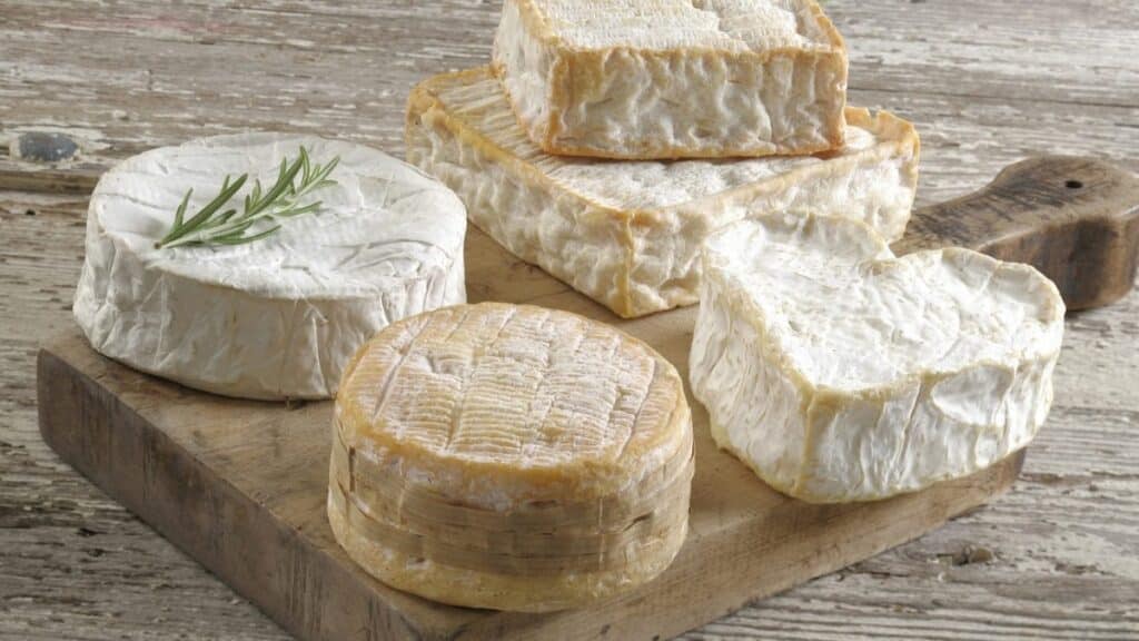 A rustic wooden board displaying an assortment of Normandy cheeses, including round, square, and heart-shaped varieties of camembert cheese, with a sprig of fresh rosemary on top of one round cheese.