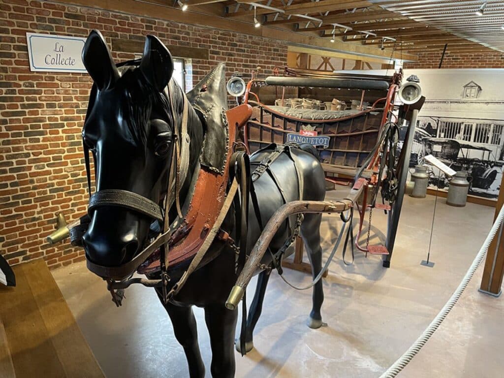 A museum exhibit featuring a black horse statue harnessed to a traditional milk cart labeled "LANQUETOT," showcasing historical camembert cheese transportation methods.