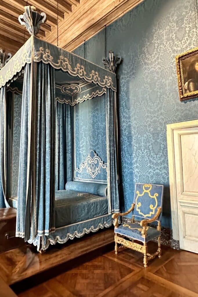An opulent bedroom in Château de Chambord, featuring a blue velvet canopy bed with intricate embroidery, matching wallpaper, and a gilded chair. The room reflects the grandeur of French Renaissance design.