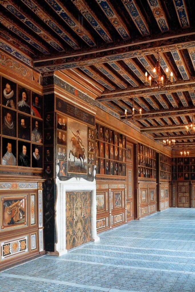A long hall inside Château de Chambord, adorned with wooden paneling, elaborate ceiling beams, and a collection of Renaissance portraits. A grand fireplace sits at the center, adding to the regal ambiance.