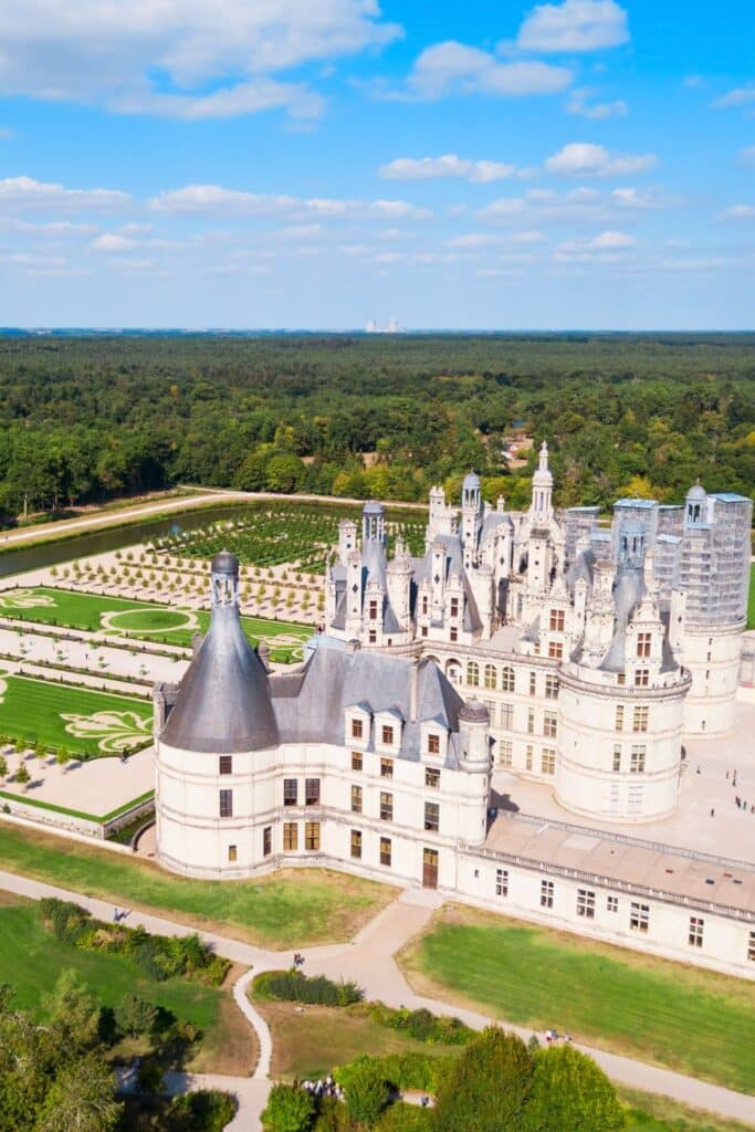 A stunning aerial perspective of Château de Chambord, showcasing its sprawling formal gardens, geometric patterns, and vast green landscape. The castle’s towers and domes rise majestically over the Loire Valley.